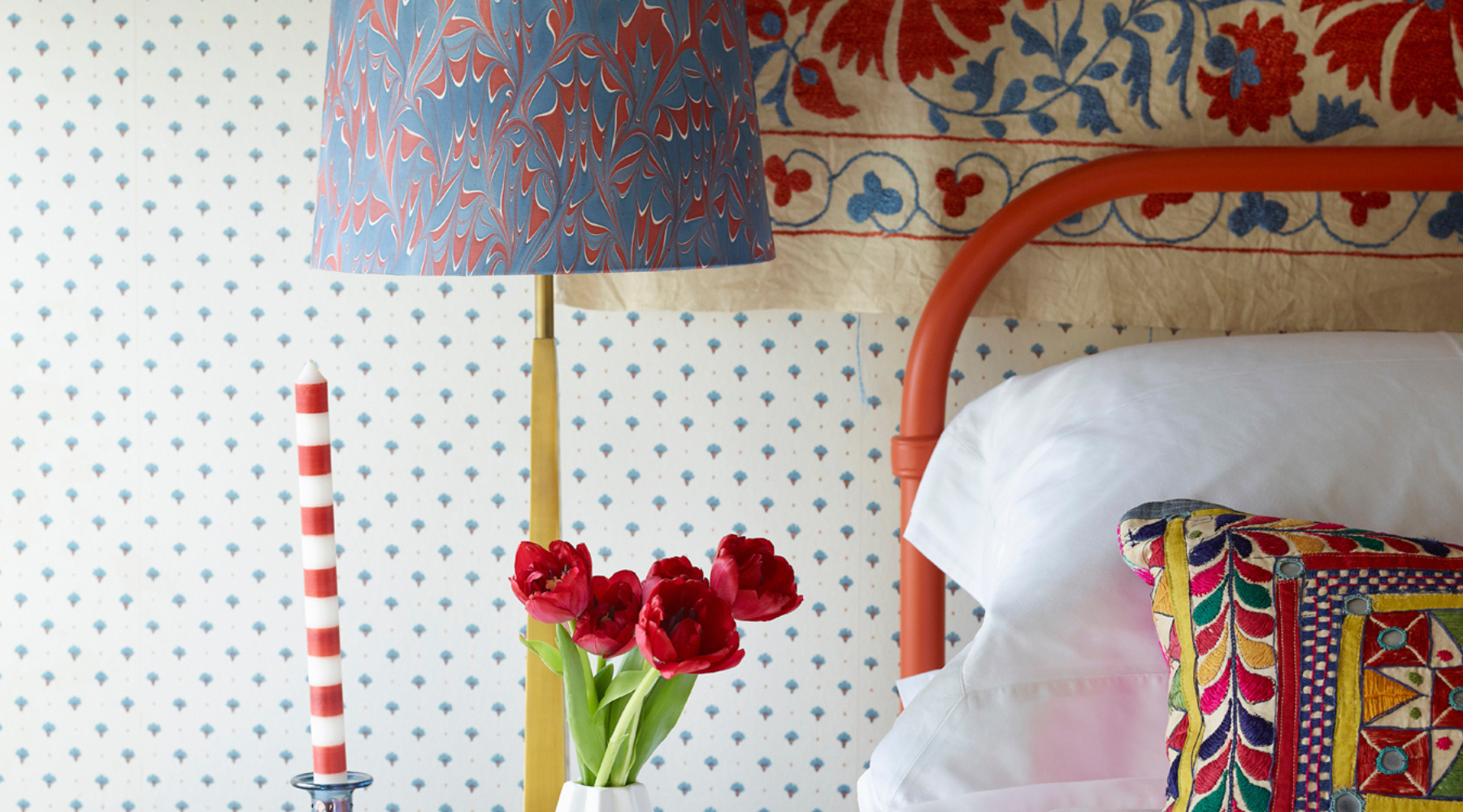 Close up of a red bed frame with brightly coloured cushions and lamp. A red and white striped candle sits on the bedside table.